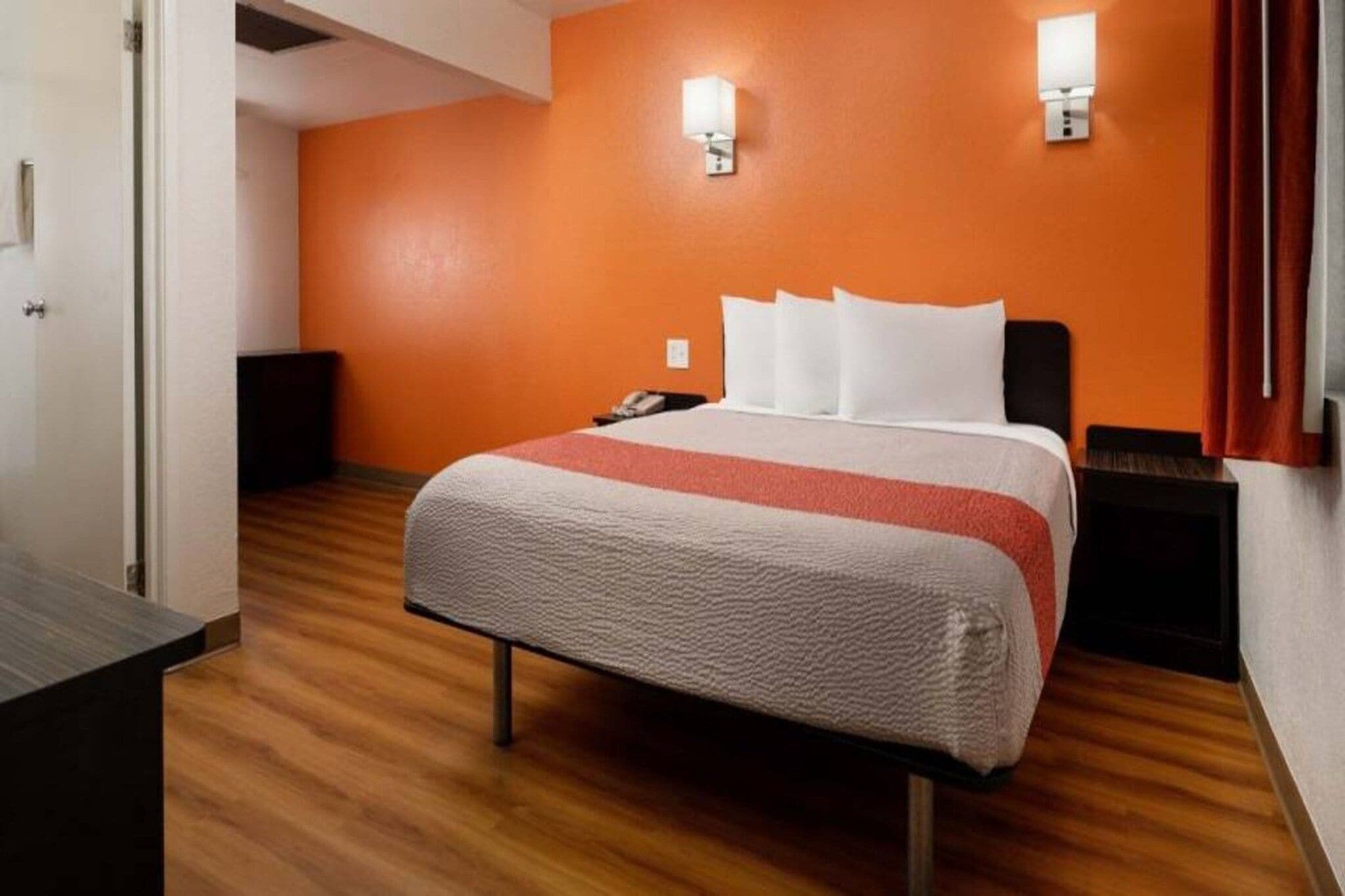 A hotel room with a bed, wooden flooring, an orange wall, and a nightstand with a lamp.