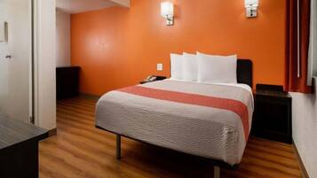 A hotel room with a bed, wooden flooring, an orange wall, and a nightstand with a lamp.