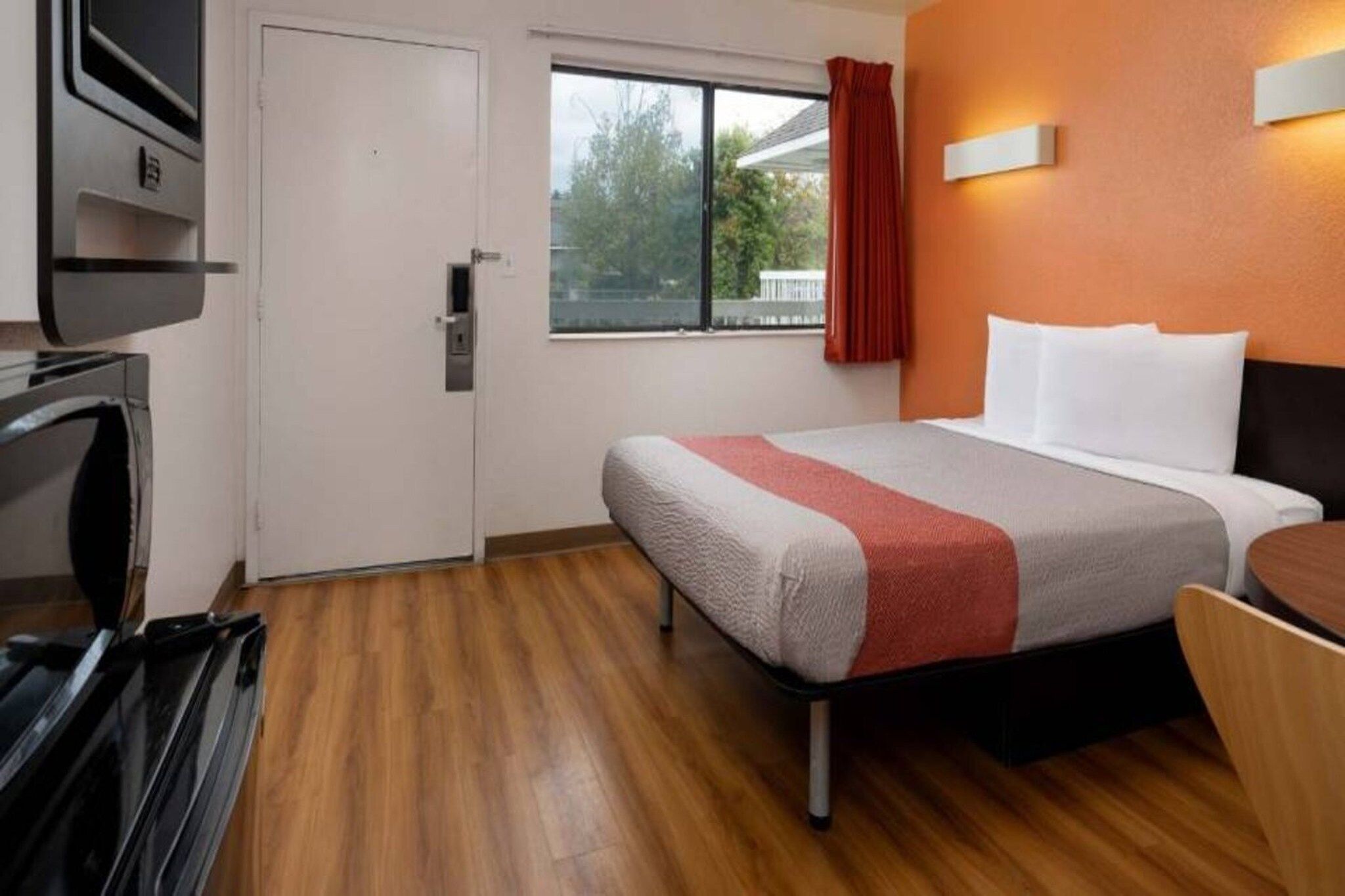 A hotel room with a bed, a window with a view of trees, a door, and a wall-mounted light fixture.
