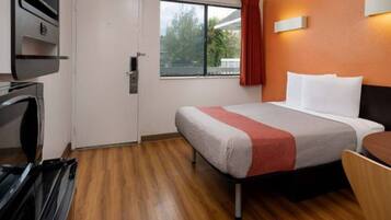 A hotel room with a bed, a window with a view of trees, a door, and a wall-mounted light fixture.
