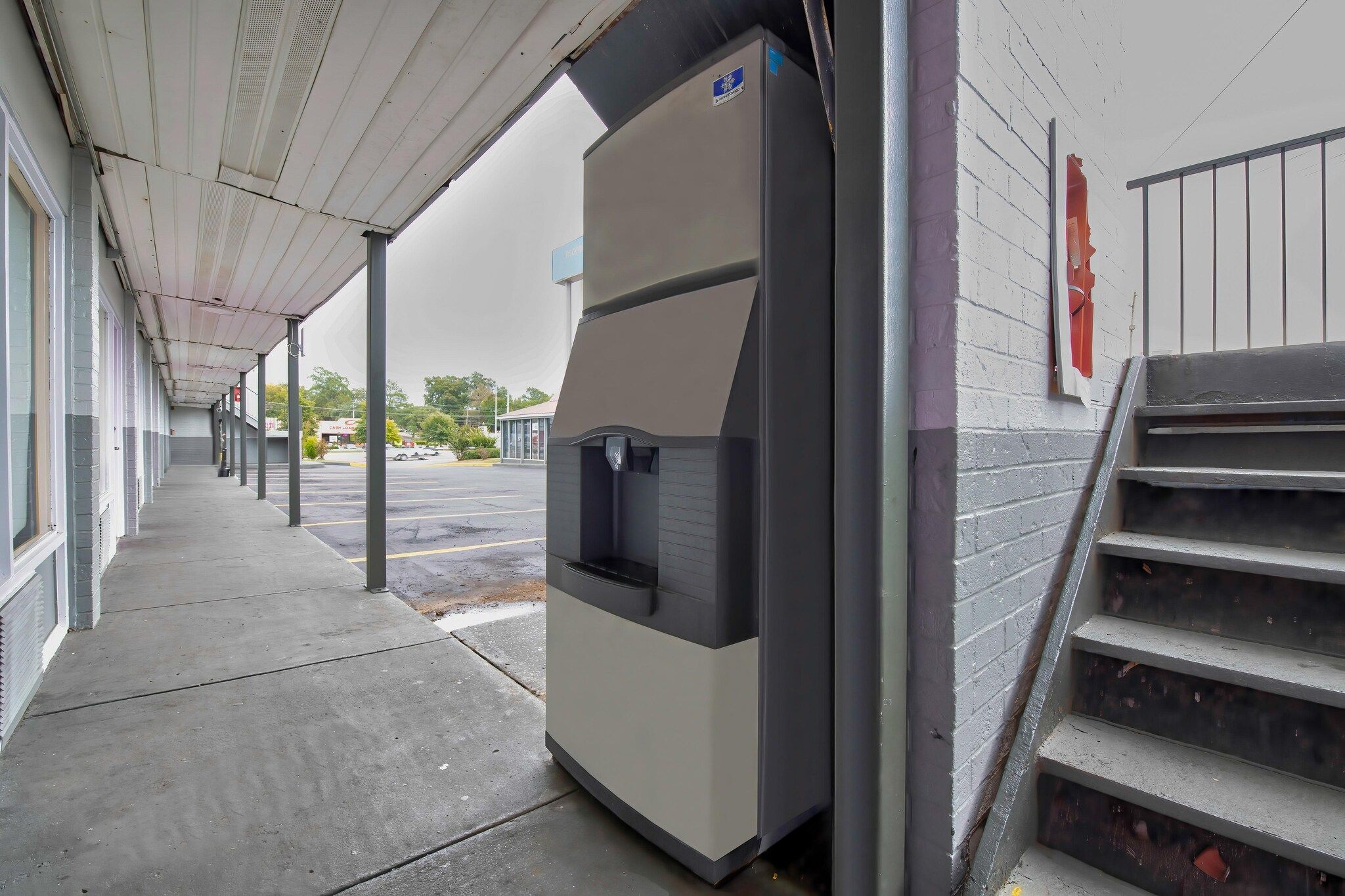 A covered walkway leading to a building with a large, modern-looking structure, possibly a vending machine or equipment, and a set of stairs leading up to a door.