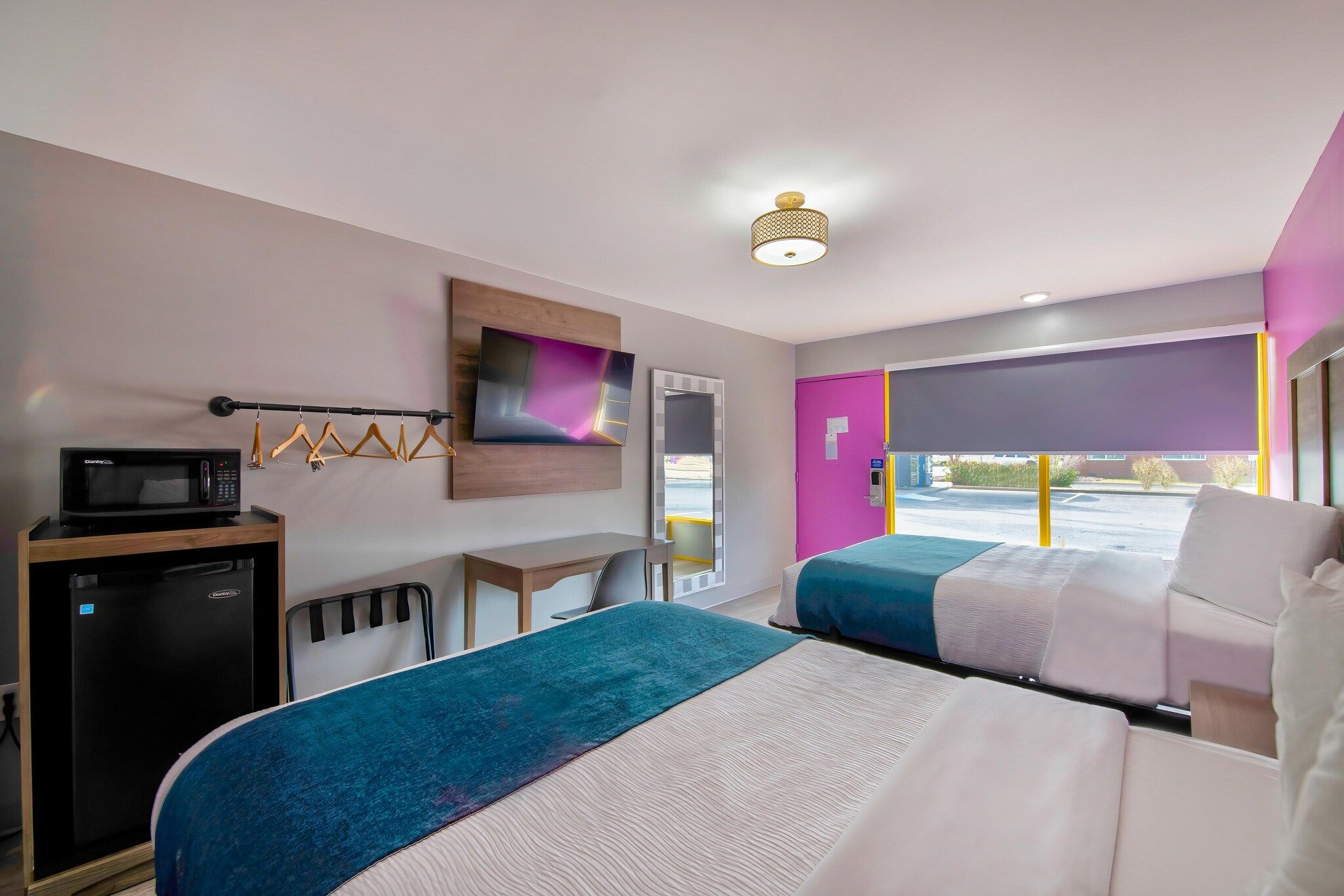A hotel room with two beds, a desk, a microwave, a refrigerator, a TV, and a window with a view of the parking lot.