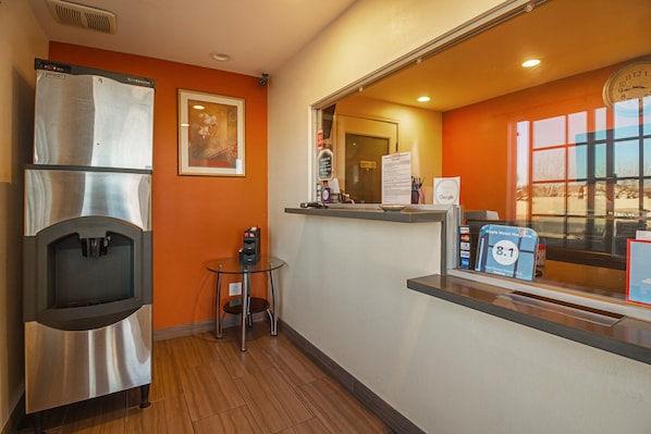 A hotel room with a minibar, a small table, a chair, a window with a view, and a reception area with a counter and a computer.