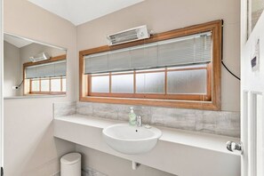 Een badkamer met een wit wastafelmeubel, een spiegel, een raam met zonwering en een wandmodel airconditioner.