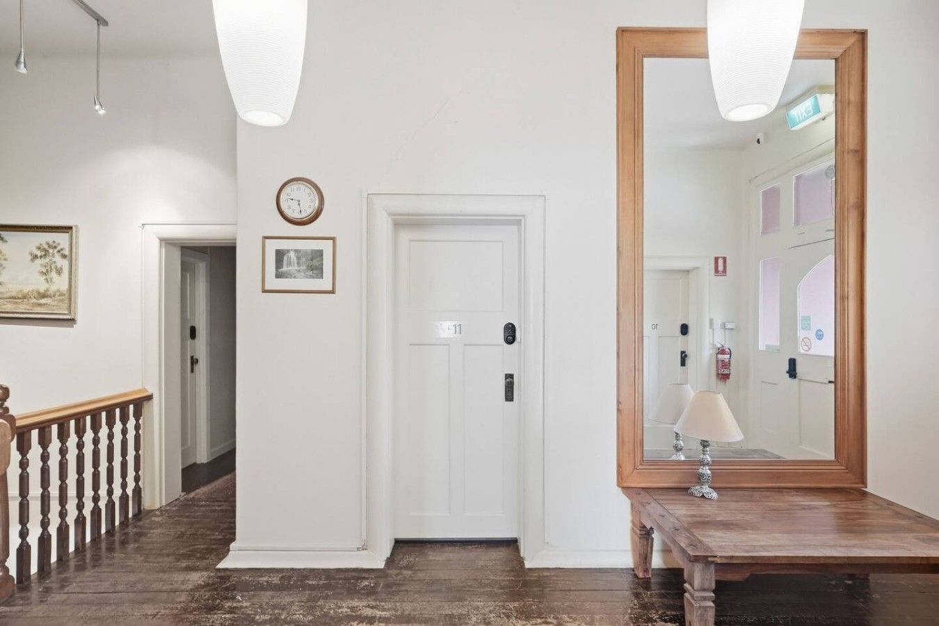 A hallway with a wooden railing, a white door with a number, a wall-mounted clock, and a mirror reflecting a door and a fire extinguisher.