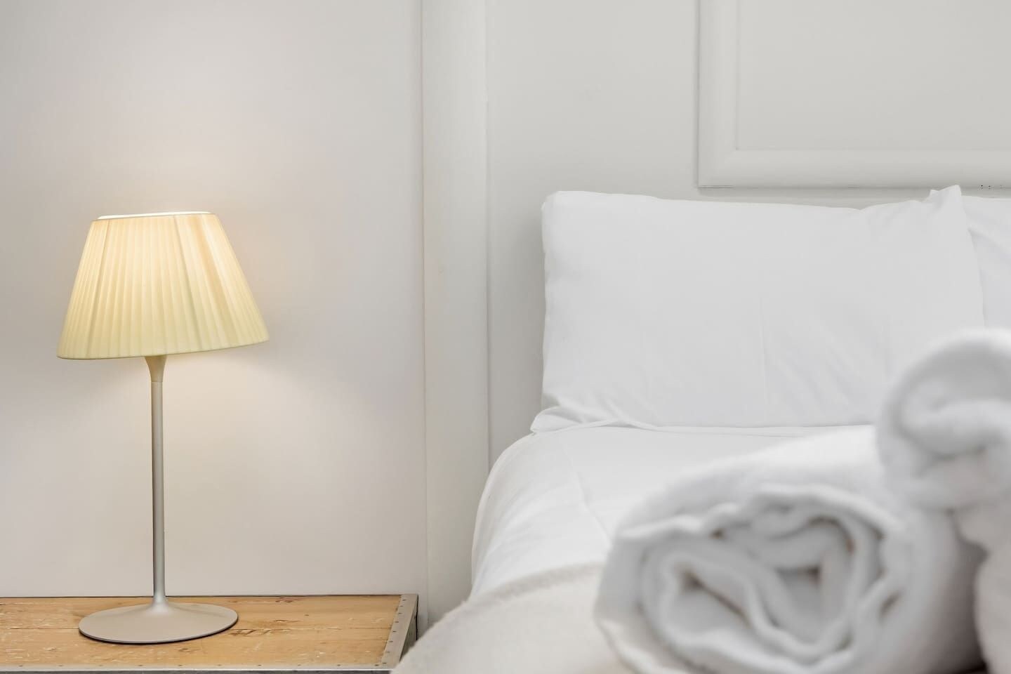 Un letto rifatto con lenzuola bianche e un asciugamano arrotolato, una lampada da comodino su un tavolino in legno e una parete semplice.