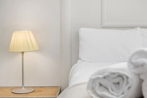 A neatly made bed with white linens and a rolled towel, a bedside lamp on a wooden nightstand, and a plain wall.