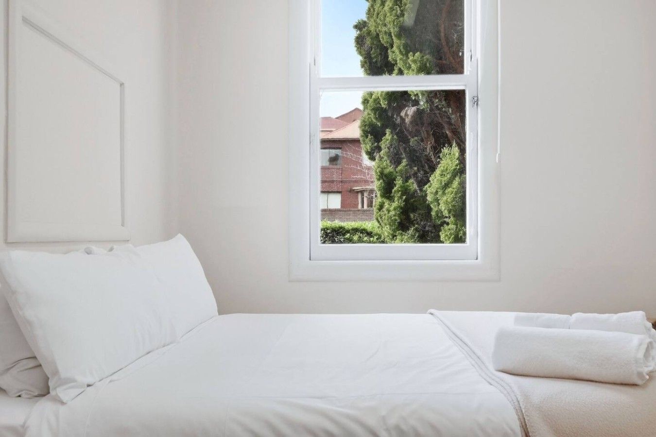 Un dormitorio con cama, ropa de cama blanca, ventana y vistas a árboles y un edificio.