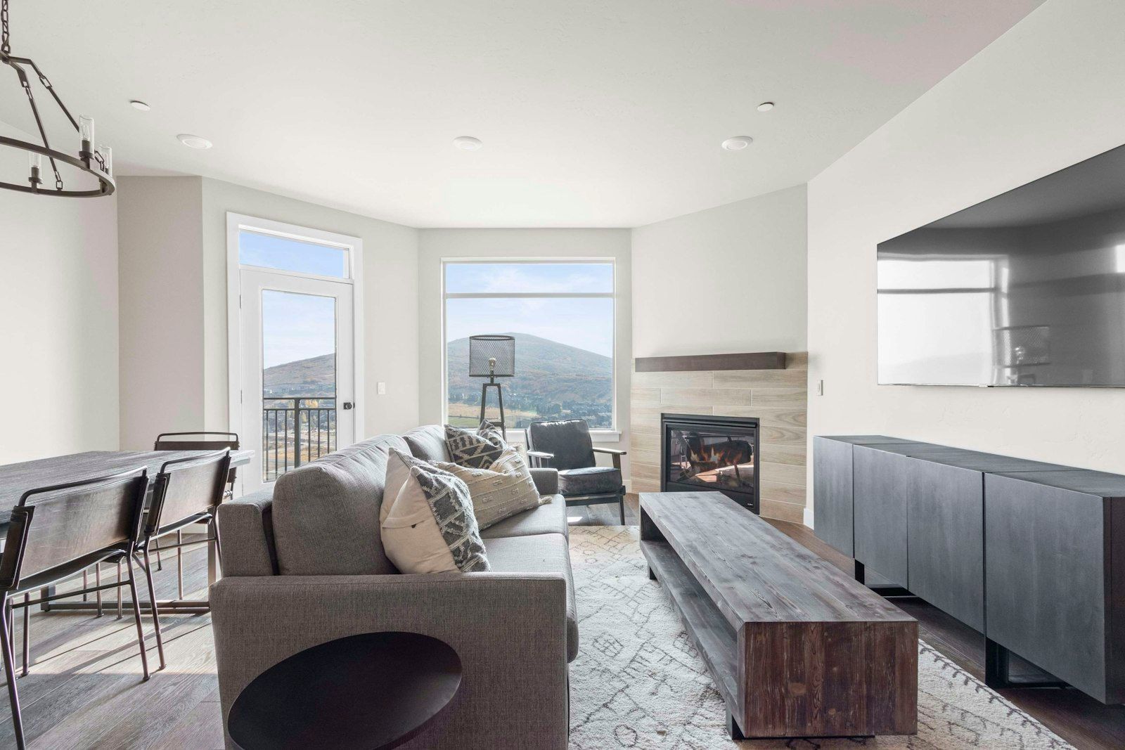 A modern living room with a fireplace, a dining area with a table and chairs, and large windows offering a view of the landscape.