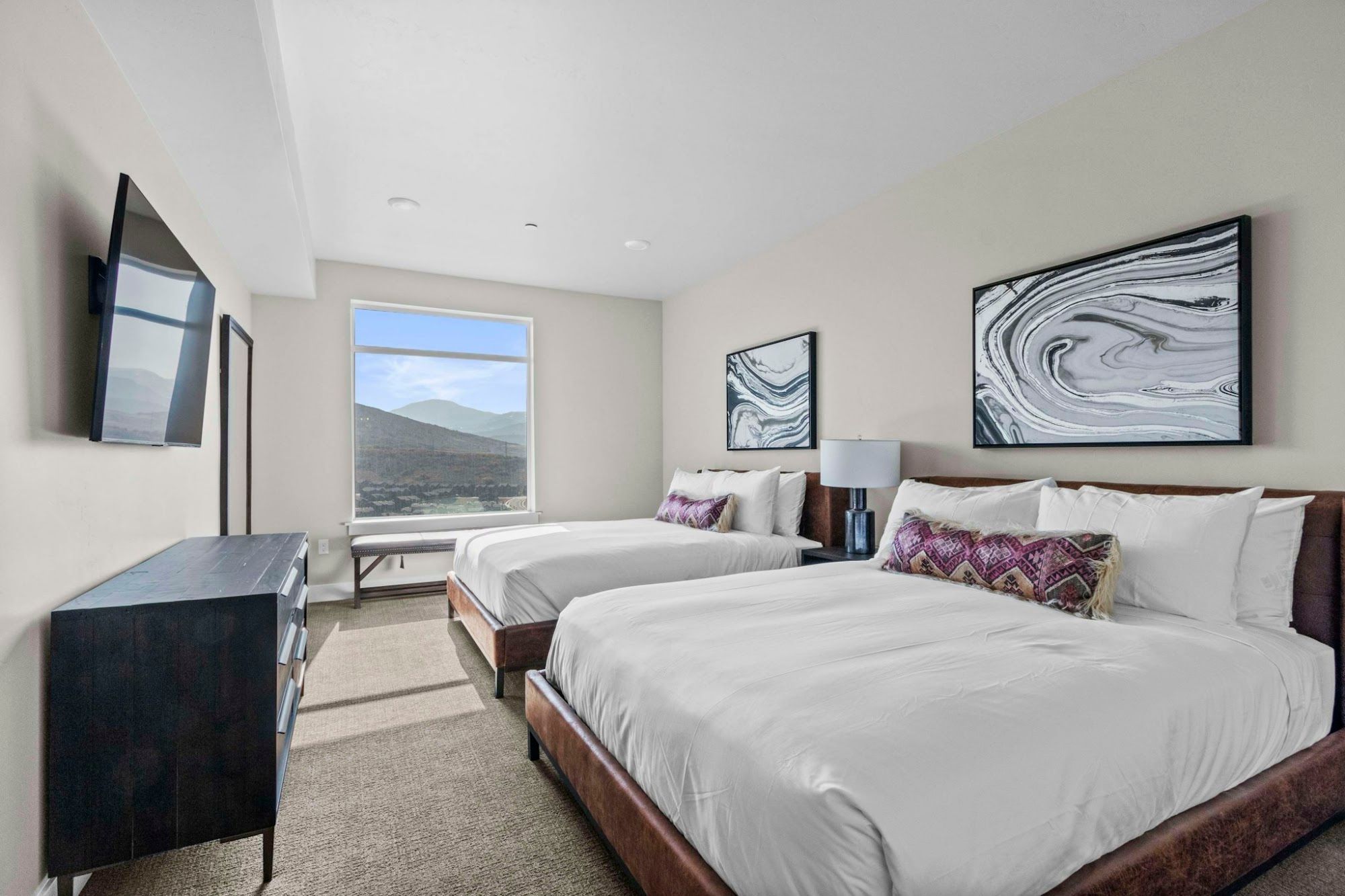 A hotel room with two beds, a desk, a TV, and artwork on the walls.