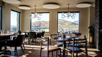 A dining area with tables and chairs, large windows with a view of hills, and a stone wall.