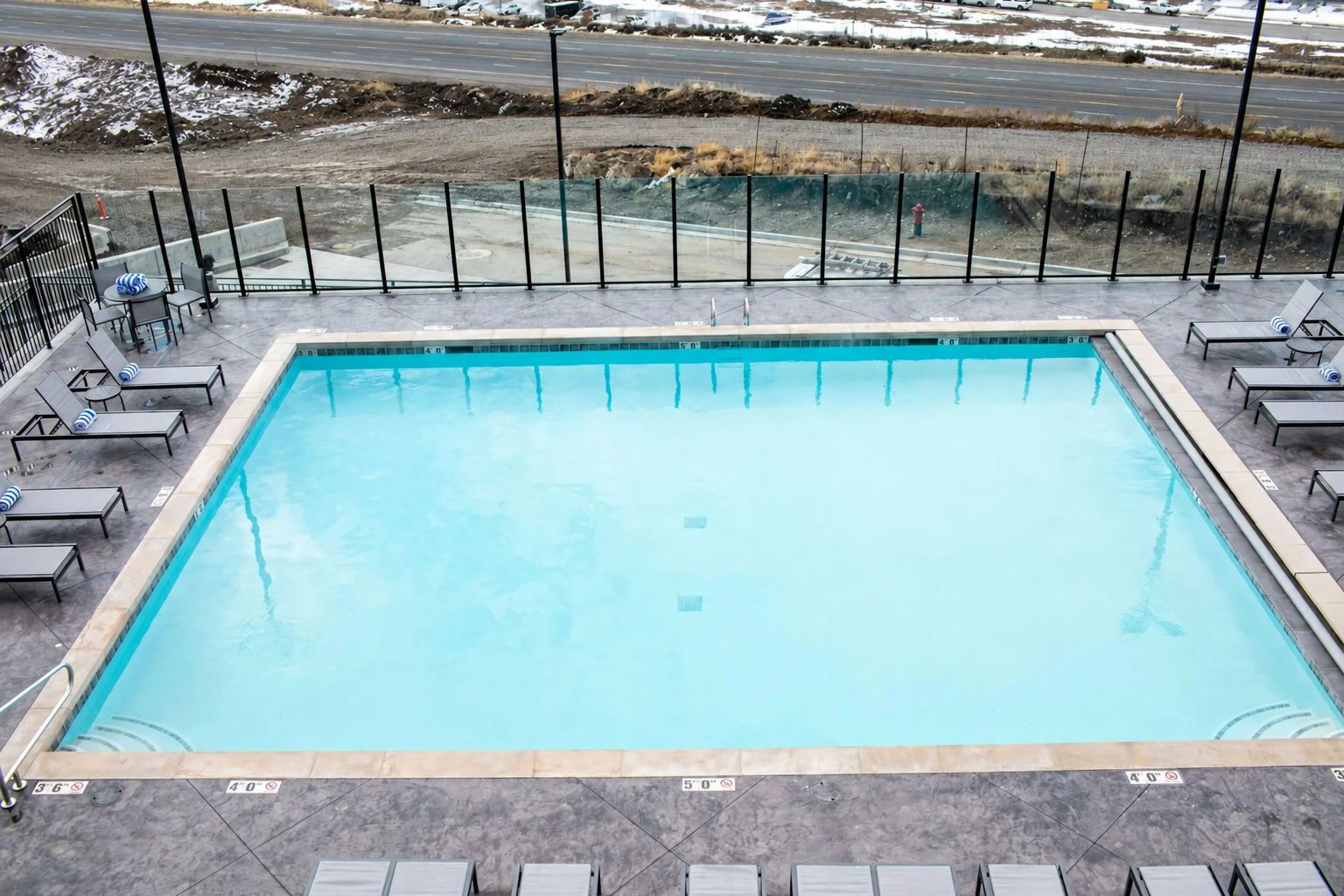 A rectangular swimming pool with a tiled deck, surrounded by lounge chairs and a metal fence.