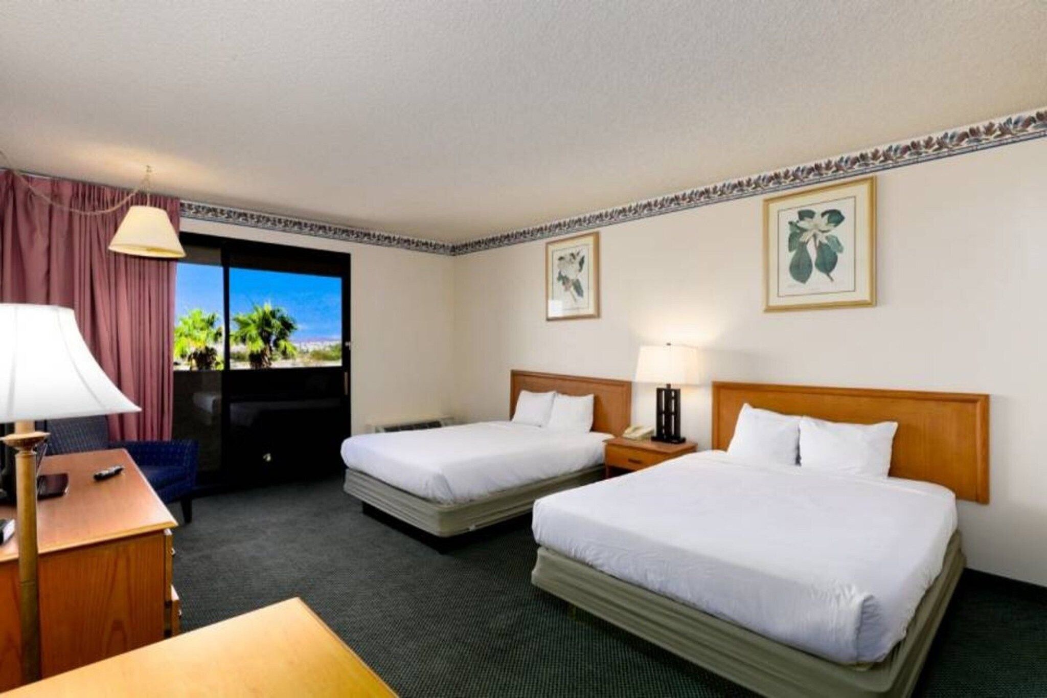 A hotel room with two beds, a desk, a chair, a lamp, and a view of palm trees through the window.