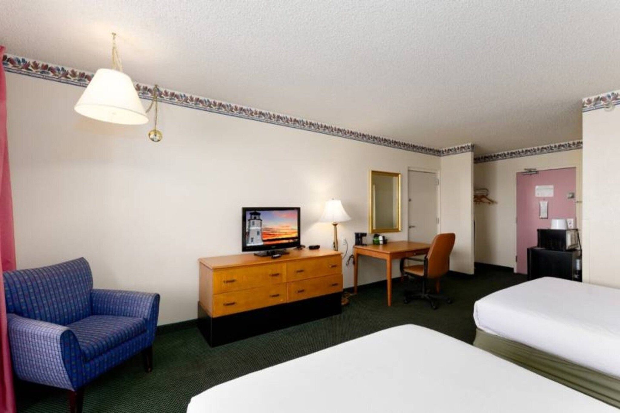 A hotel room with two beds, a desk, a chair, a dresser, a television, and a lamp.