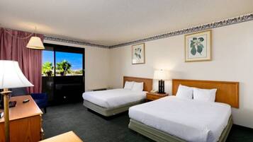 A hotel room with two beds, a desk, a chair, a lamp, and a view of palm trees through the window.