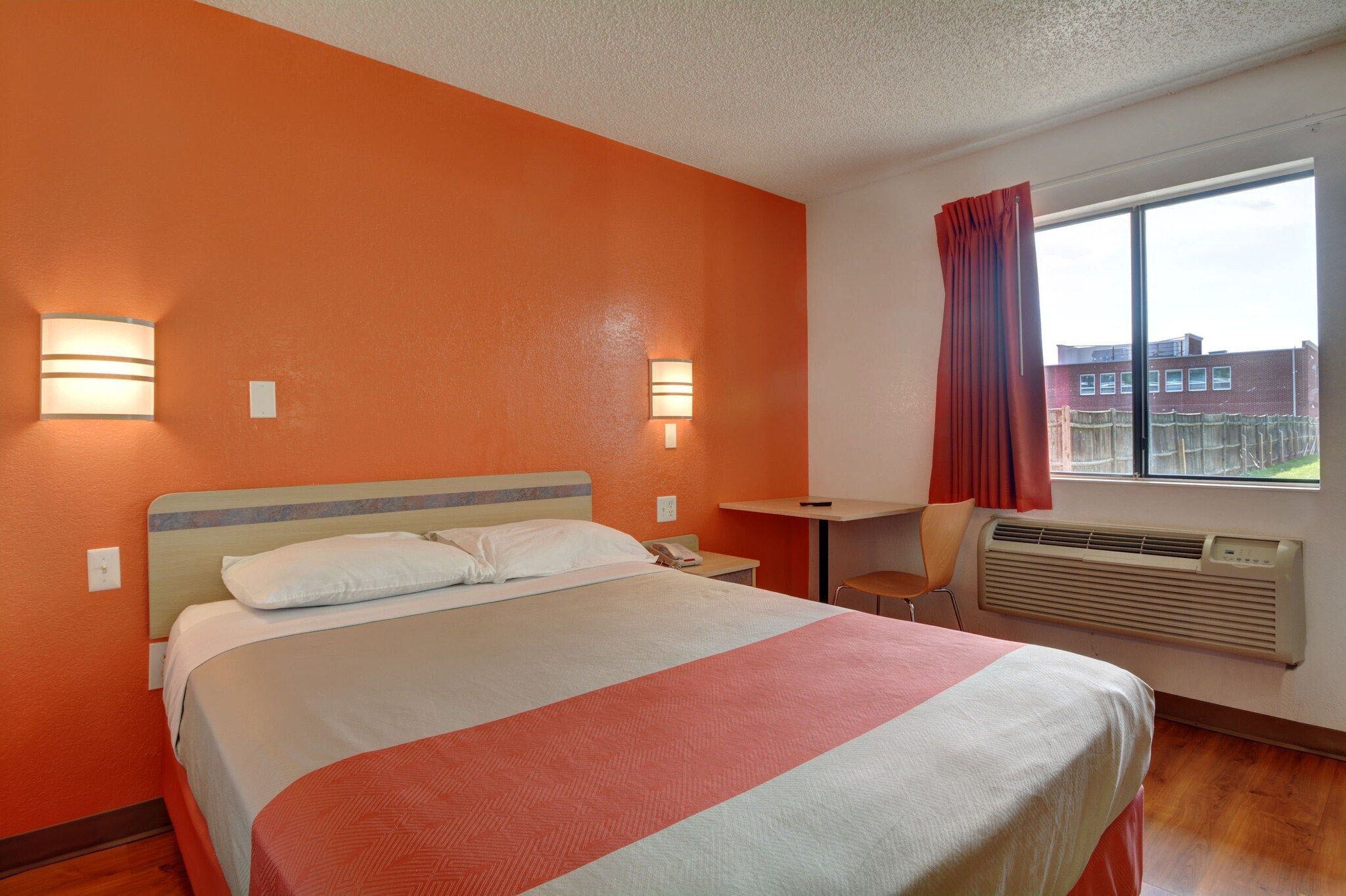 A hotel room with a bed, bedside tables, a desk, a chair, a window with curtains, and an air conditioning unit.