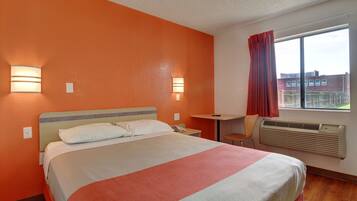 A hotel room with a bed, bedside tables, a desk, a chair, a window with curtains, and an air conditioning unit.