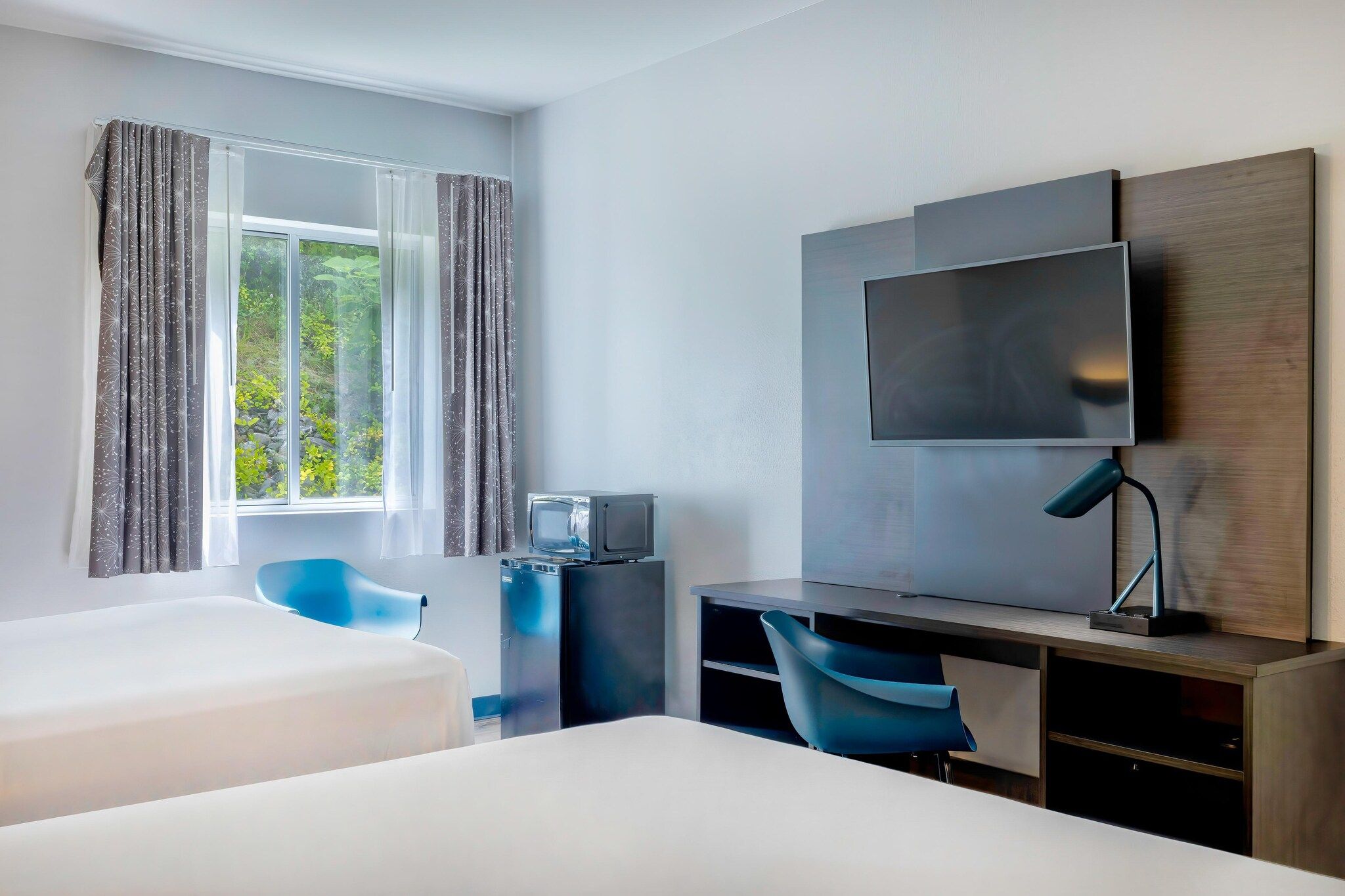 Una habitación de hotel con cama, escritorio con silla, televisor y ventana con cortinas.