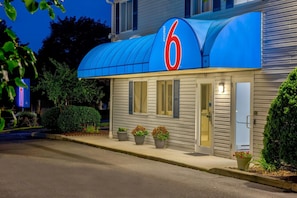 Un motel con un llamativo letrero con el número '6', un acceso cubierto y plantas en macetas a la entrada.