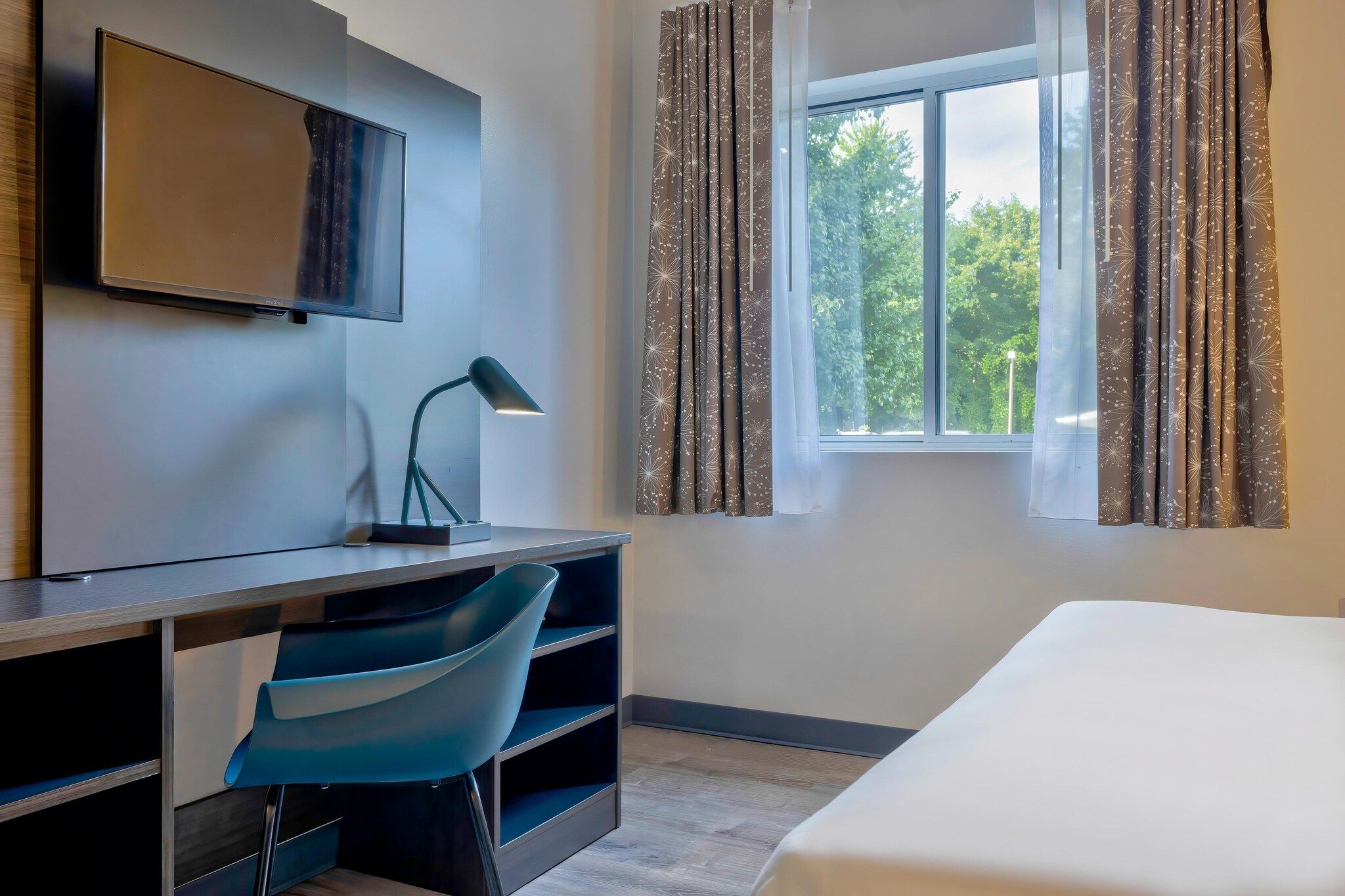 A hotel room with a bed, a desk with a chair, a TV, and a window with curtains.