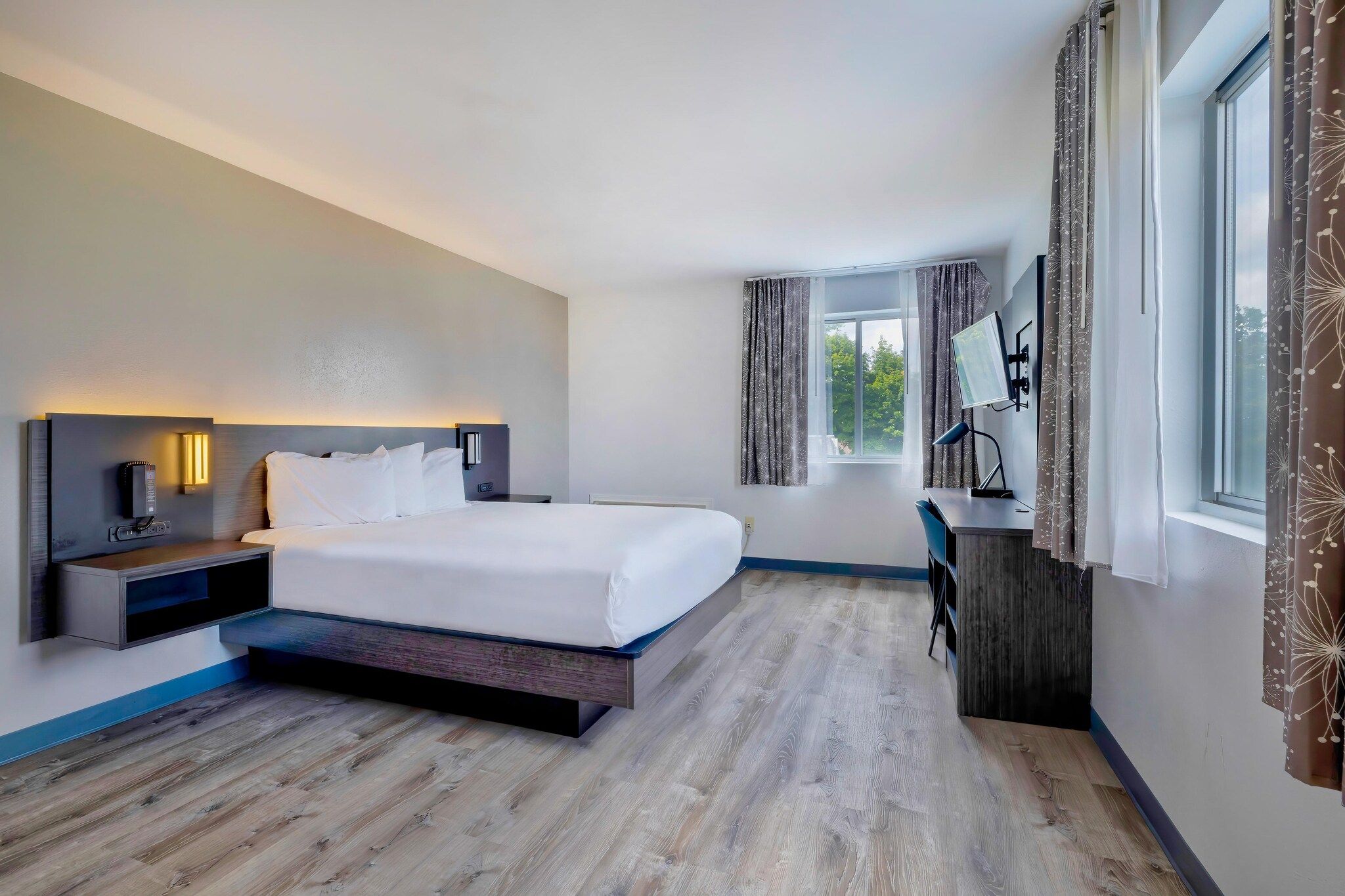 A modern hotel room with a large bed, bedside tables, a desk with a chair, a flat-screen TV, and a window with curtains.