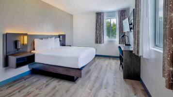 A modern hotel room with a bed, bedside table, lamp, desk, and window.