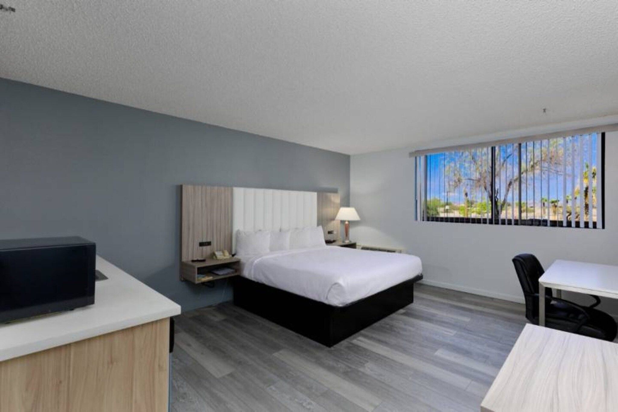 Een hotelkamer met een bed, nachtkastje, lamp, televisie, bureau, stoel en een raam met uitzicht.