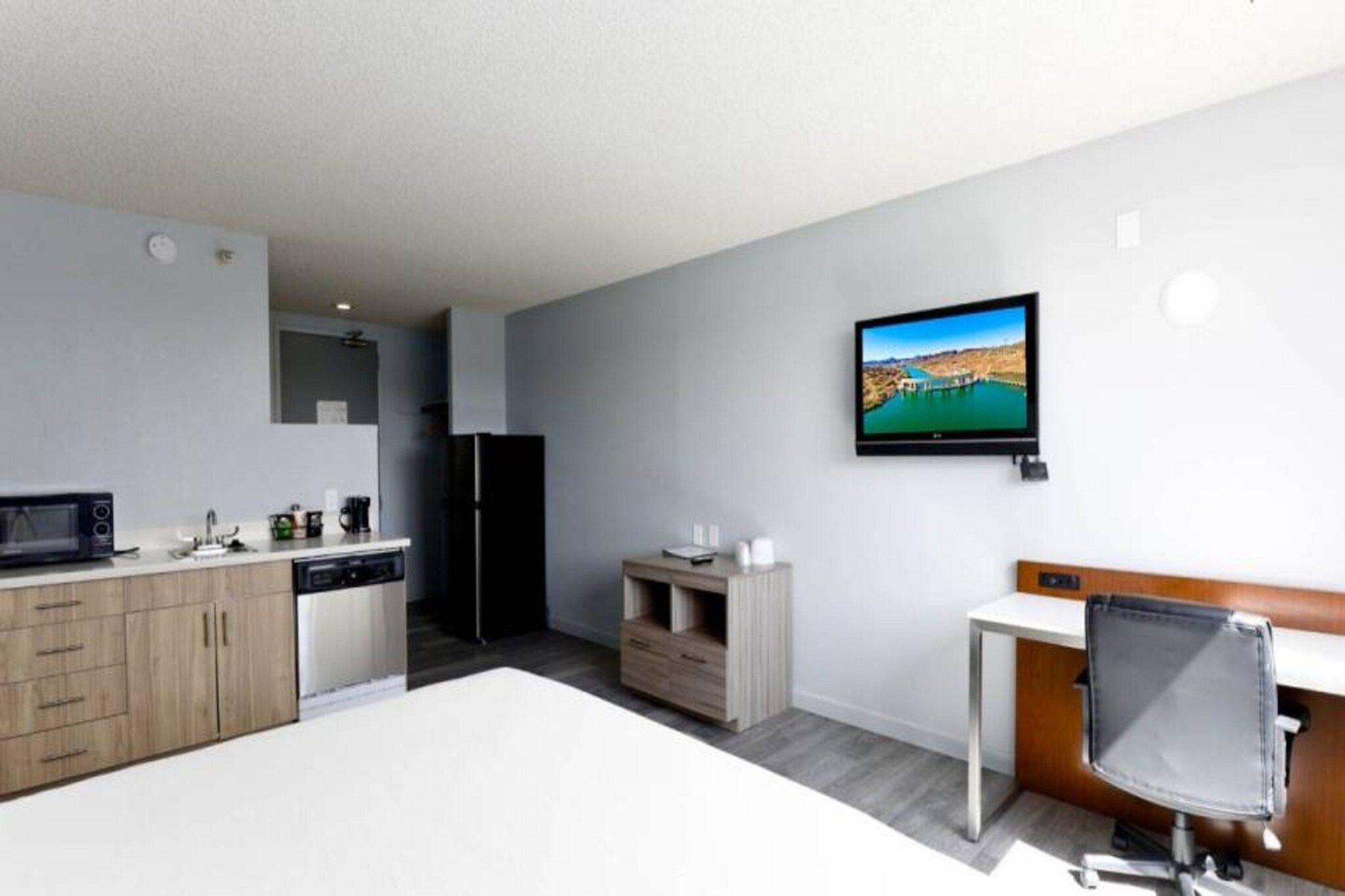 A modern hotel room with a kitchenette, a flat-screen TV mounted on the wall, and a desk with a chair.