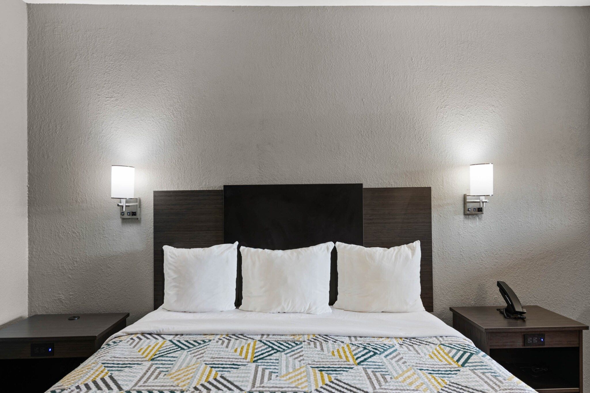 A hotel room with a bed, two bedside tables, a wall-mounted lamp, and a telephone.