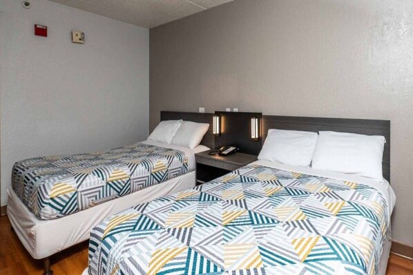 A hotel room with two beds, each with patterned bedding, a bedside table with a lamp, and a wall-mounted phone.