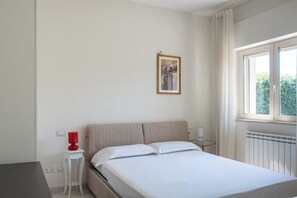 Ett hotellrum med en säng, ett nattduksbord, en röd lampa, en inramad tavla på väggen och ett fönster med gardiner.