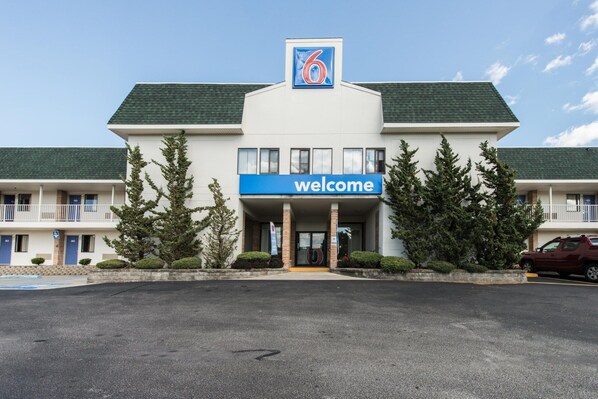 Ein Motel mit einem 'Welcome'-Schild, einem Parkplatz und einem roten Auto vor der Tür.