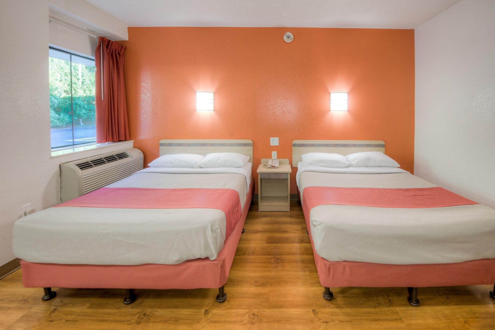 Una camera d'albergo con due letti, una finestra con tende, un condizionatore d'aria e un lampadario a parete.