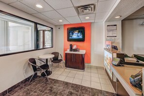 Zona de recepción de hotel con un televisor montado en la pared, una pequeña recepción con una computadora y un área de descanso con dos sillas y una mesa.