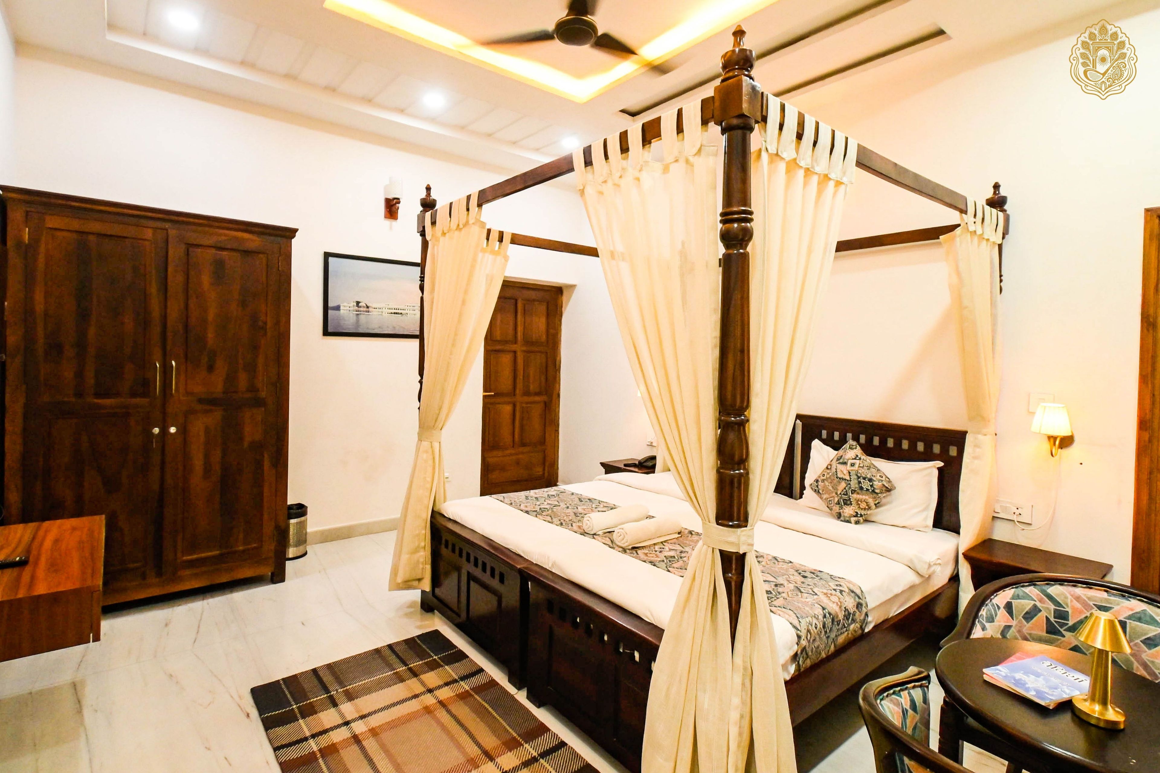A four-poster canopy bed with a wooden frame and white curtains, a wooden wardrobe, a small bedside table with a lamp, and a patterned rug.