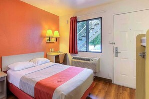 A hotel room with a bed, a desk, a chair, a window with curtains, and an air conditioning unit. - 1 Full Bed | ADA/RI Shower (Pinole)