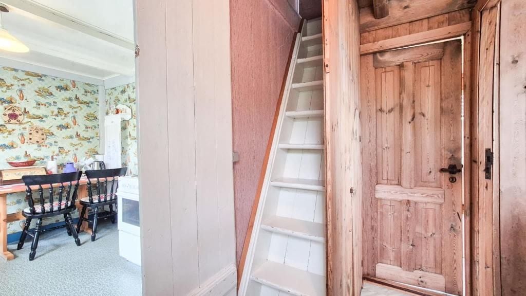 A wooden staircase leading to a door, a dining area with a table and chairs, and a wall with a patterned wallpaper.