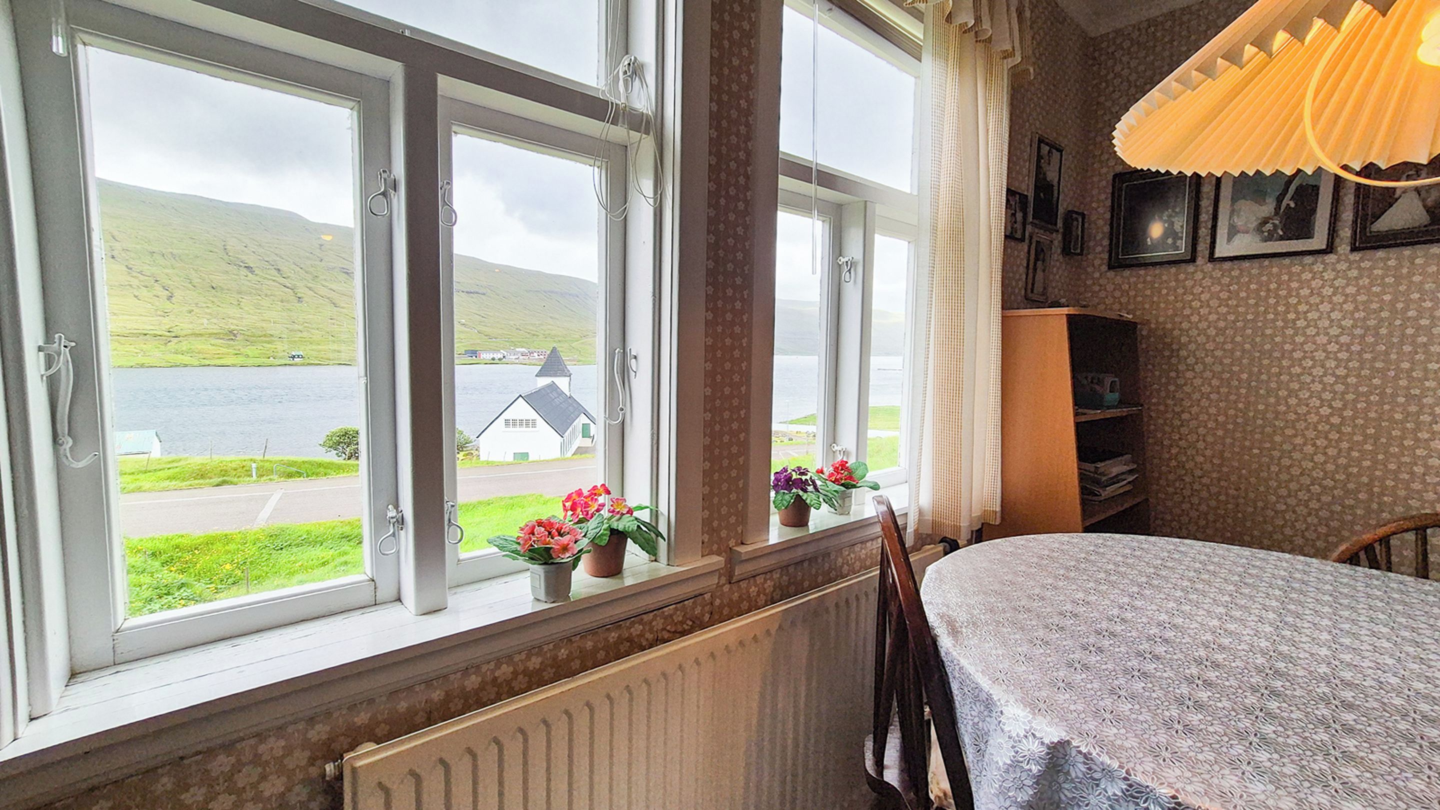 A room with a view of a lake and a white building, a table with a tablecloth, and a bookshelf.