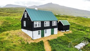 A black and white house with a green roof, surrounded by grassy hills and mountains in the background.