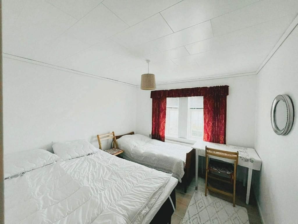Una habitación con dos camas individuales, un escritorio con silla, una ventana con cortinas rojas y una lámpara colgante.