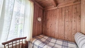 A wooden cabin room with a bed, a window with curtains, and a wall-mounted light fixture.