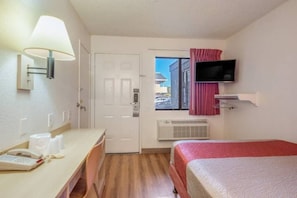 A hotel room with a bed, a desk with a telephone, a lamp, an air conditioning unit, and a television mounted on the wall. - Spacious 1 Queen Bed | ADA/Tub (Fairfield)