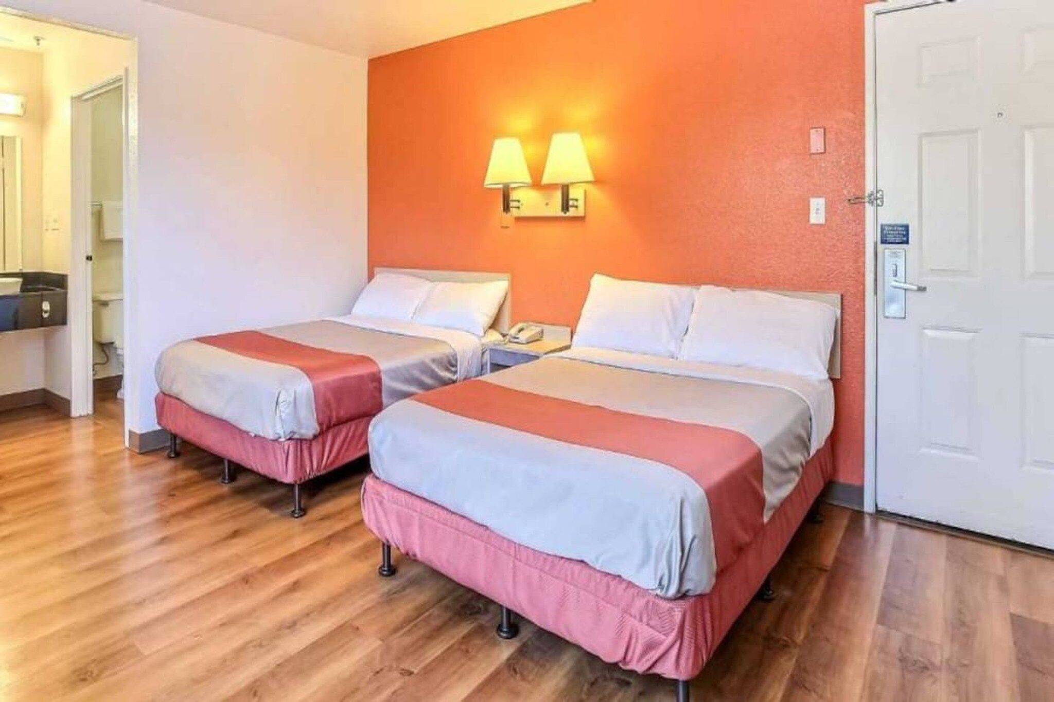 Une chambre d’hôtel avec deux lits, un plancher en bois, une porte blanche et une lampe fixée au mur.