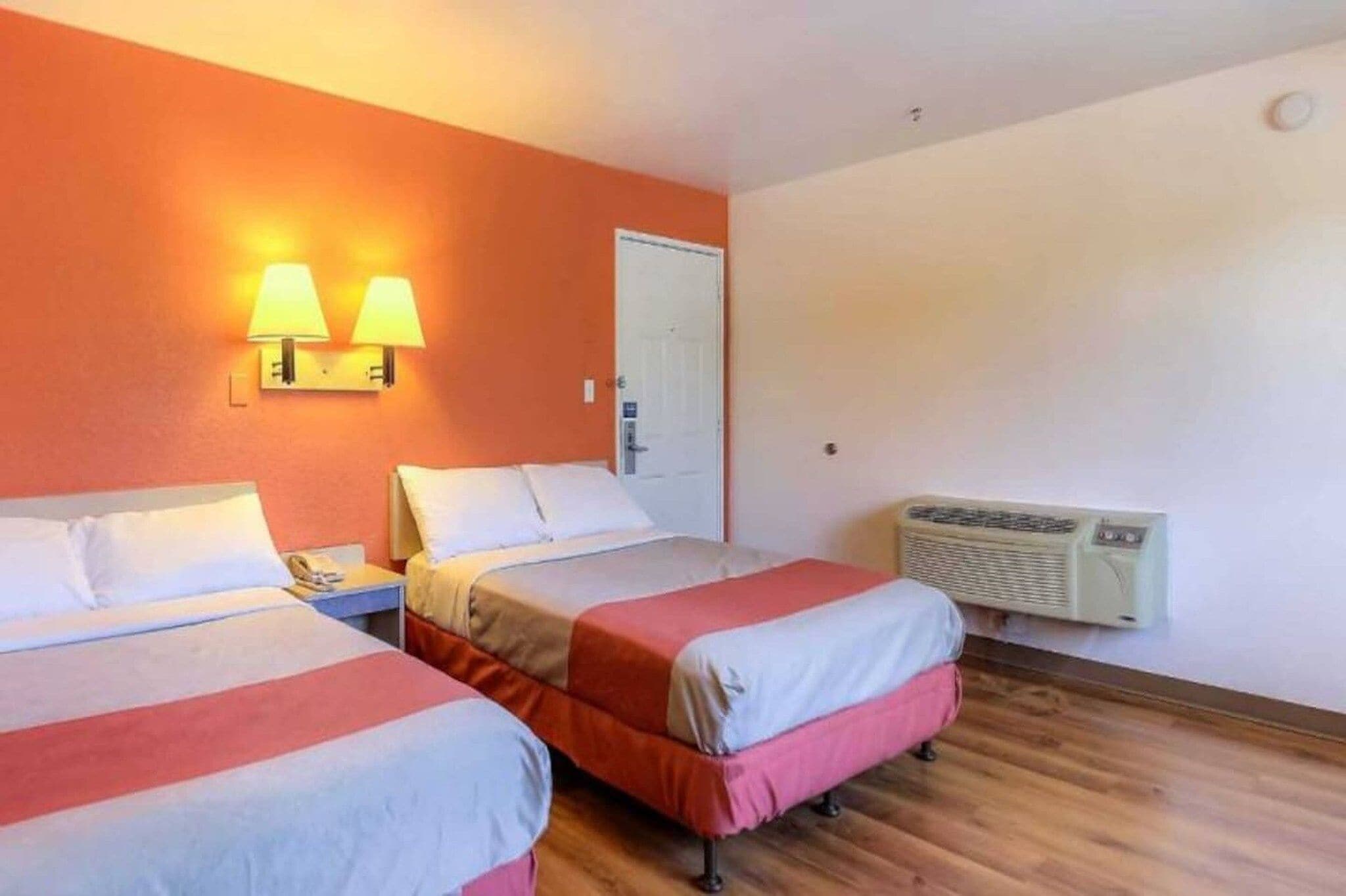 A hotel room with two beds, a nightstand, a lamp, an air conditioning unit, and a door.