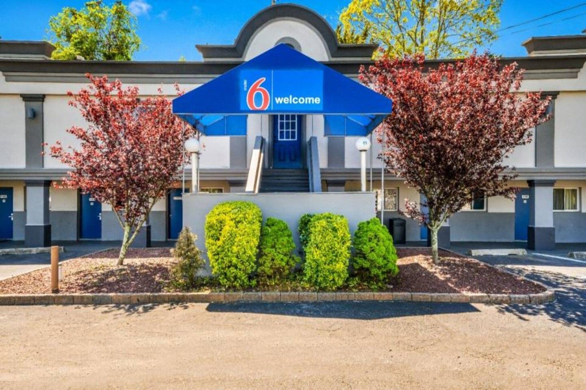 Ein Hotel Eingang mit einem blauen Schild mit der Aufschrift '6 welcome', einer Treppe, die zum Eingang führt, und zwei Bäume mit roten Blättern.