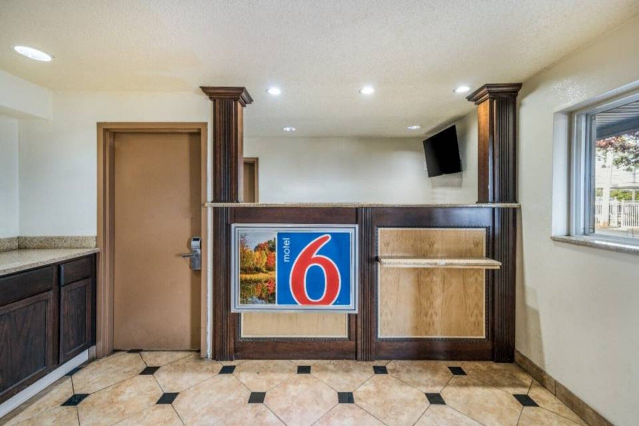 Een hotel receptie met een groot bord met het nummer 6, een houten receptiebalie, een deur en een raam met uitzicht.