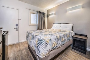 Un dormitorio con cama, mesita de noche, ventana con cortinas, puerta y lámpara fijada a la pared.