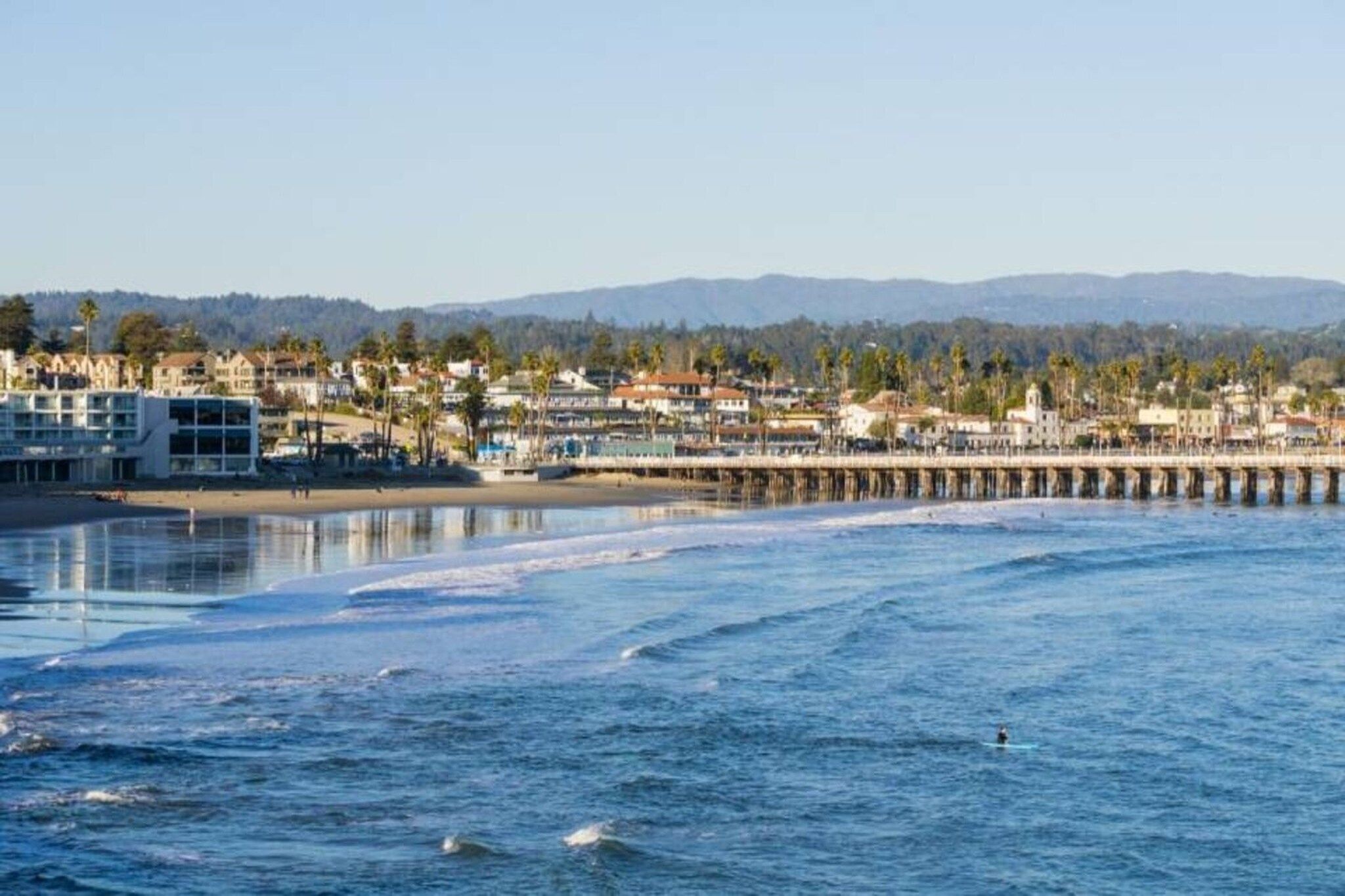 A coastal town with a pier extending into the sea, buildings along the shore, and mountains in the background.