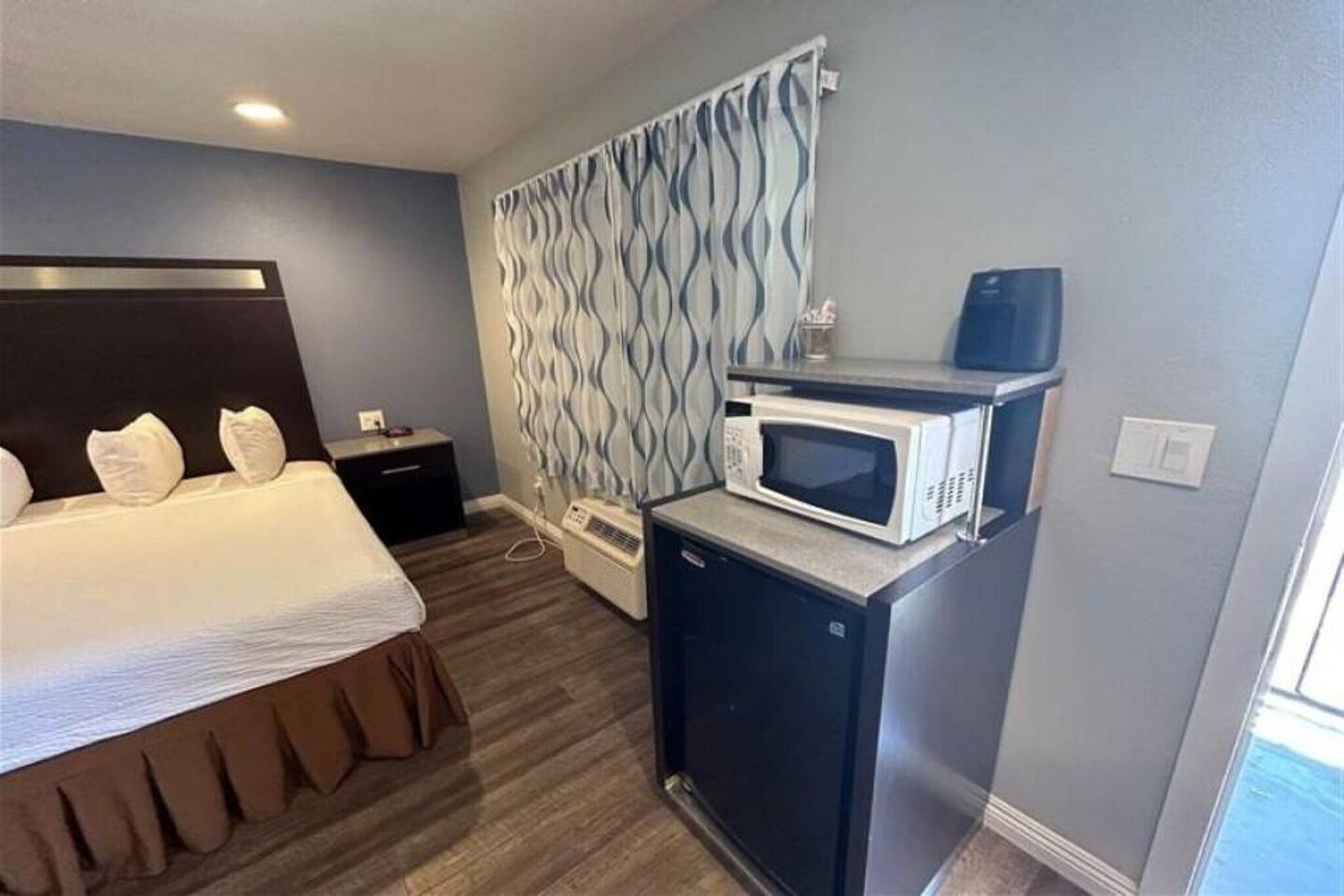 A hotel room with a bed, a bedside table, a microwave, and a wall-mounted air conditioning unit.