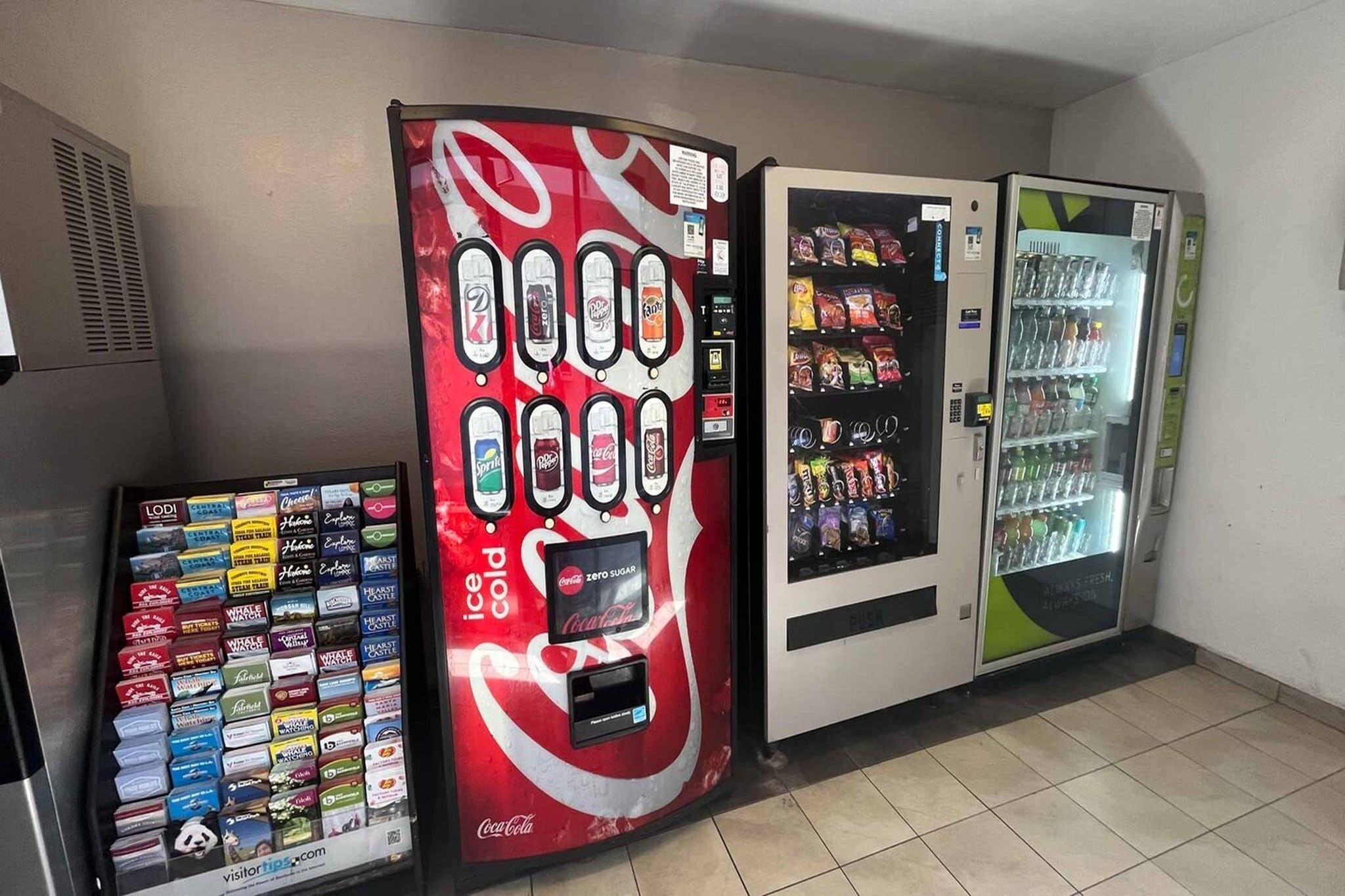 Un distributeur automatique proposant divers en-cas et boissons, un distributeur arborant la marque Coca-Cola, et un réfrigérateur contenant des bouteilles d’eau.